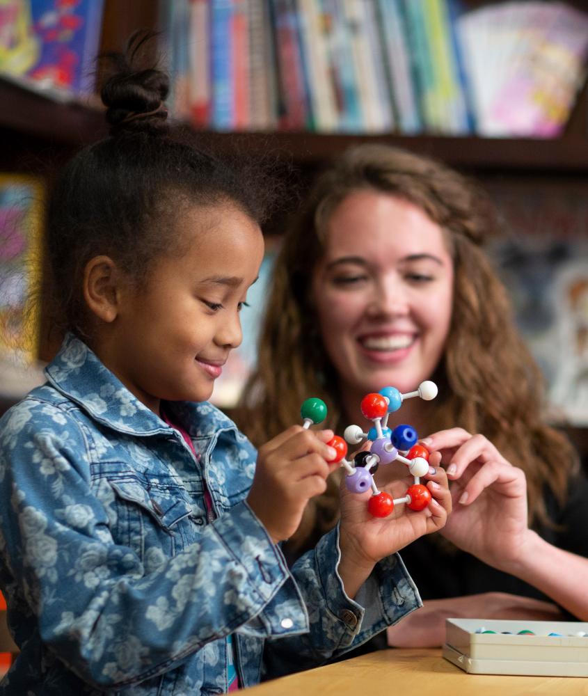 A young female teacher demonstrates and plays with chemistry molecule toy with a female student. 
