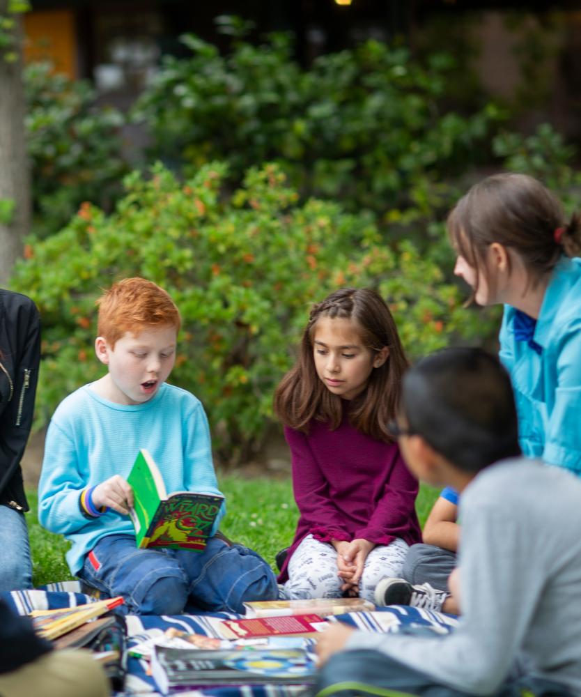 A group of elementary school students sitting outside on a blanket in circle surrounded by books. A red-haired boy in blue shirt is reading aloud from "The Wizard of Oz"