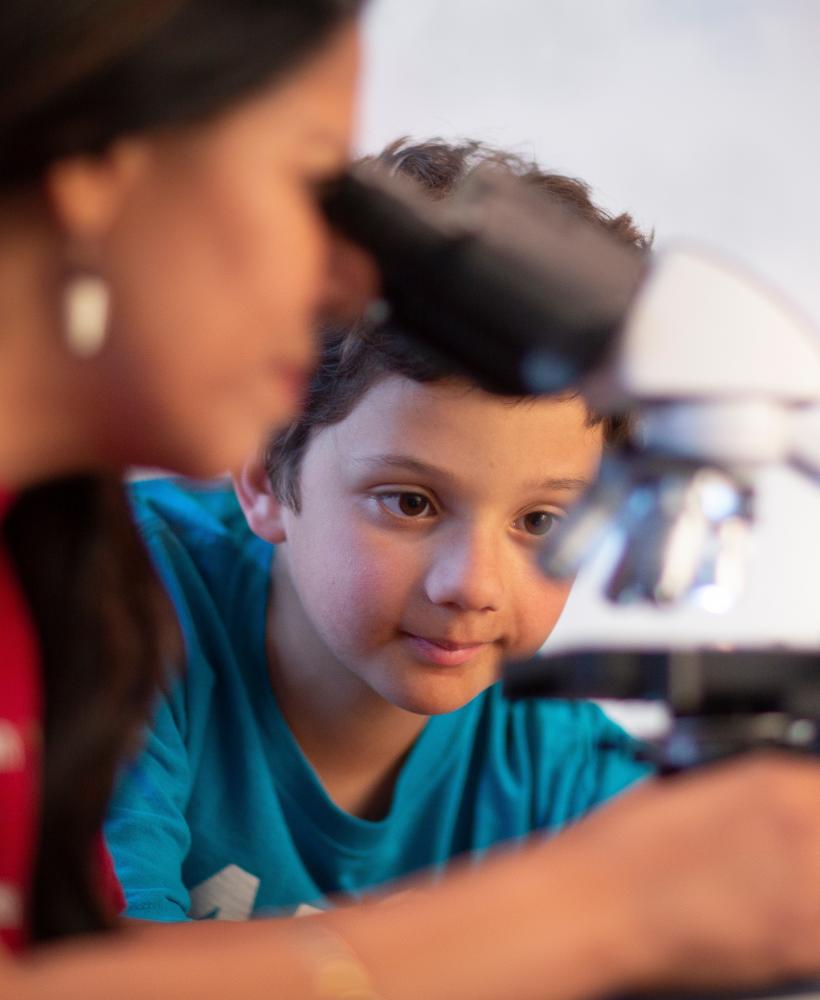 A young boy wearing a blue shirt waits for his turn to use a microscope while his teacher demonstrates how to use it. 