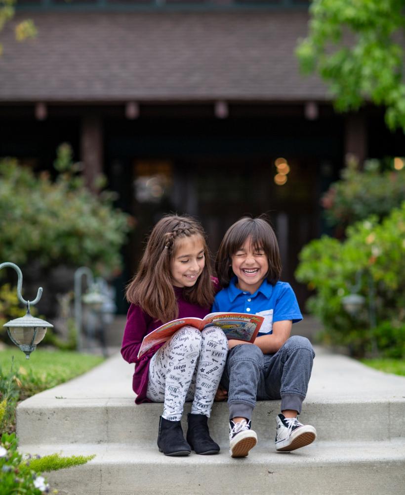 Two elementary-age students sit outdoors on a concrete step reading books and laughing. 